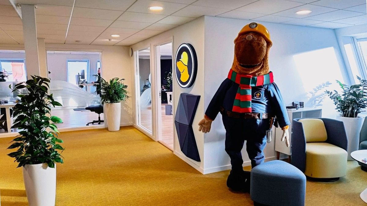 Mullvad mole mascot wearing a hard hat and scarf in the modern Mullvad VPN office with yellow carpet, plants, and circular wall logo.