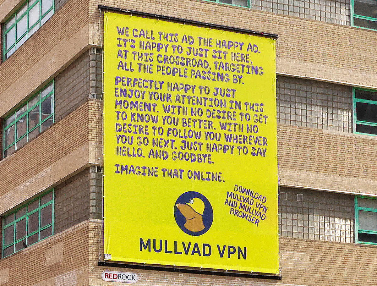 Yellow Mullvad VPN billboard on a brick building. Text: WE CALL THIS AD THE HAPPY AD. IT'S HAPPY TO JUST SIT HERE, AT THIS CROSSROAD, TARGETING ALL THE PEOPLE PASSING BY. PERFECTLY HAPPY TO JUST ENJOY YOUR ATTENTION IN THIS MOMENT. WITH NO DESIRE TO GET TO KNOW YOU BETTER. WITH NO DESIRE TO FOLLOW YOU WHEREVER YOU GO NEXT. JUST HAPPY TO SAY HELLO. AND GOODBYE. IMAGINE THAT ONLINE. DOWNLOAD MULLVAD VPN AND MULLVAD BROWSER. MULLVAD VPN.